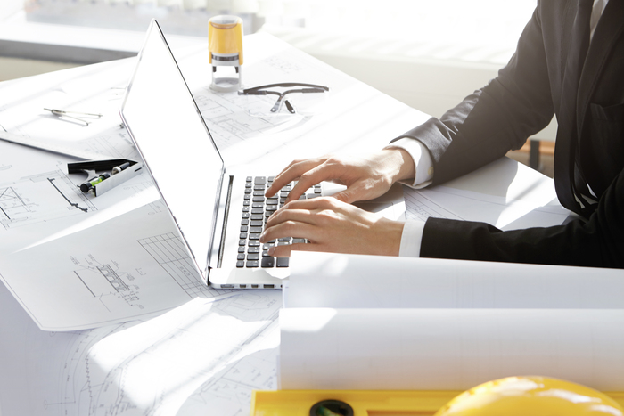 Cropped shot of architect or engineer in formal wear sitting at table with architectural project, blueprints, ruler, divider compass, typing on laptop with blank copy space screen for your content